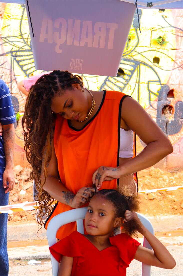 Mulher trança o cabelo de uma menina em ação de autoestima na comunidade.