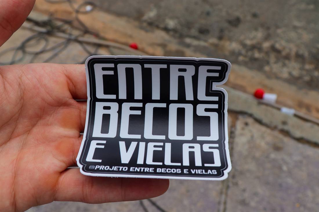Mão segura um adesivo com a marca do projeto Entre Becos e Vielas.